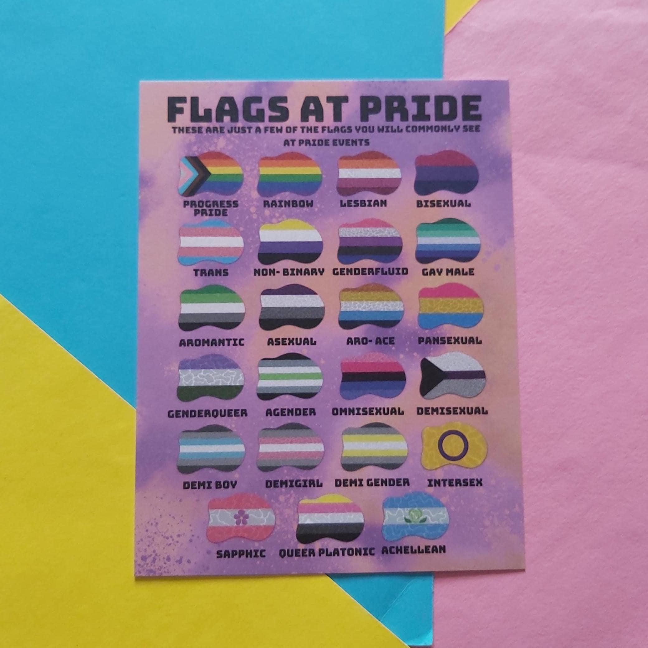 A print titled "FLAGS AT PRIDE" showing multiple small LGBTQ+ pride flags arranged in a grid on a pastel purple background, placed on a colorful pastel paper backdrop.