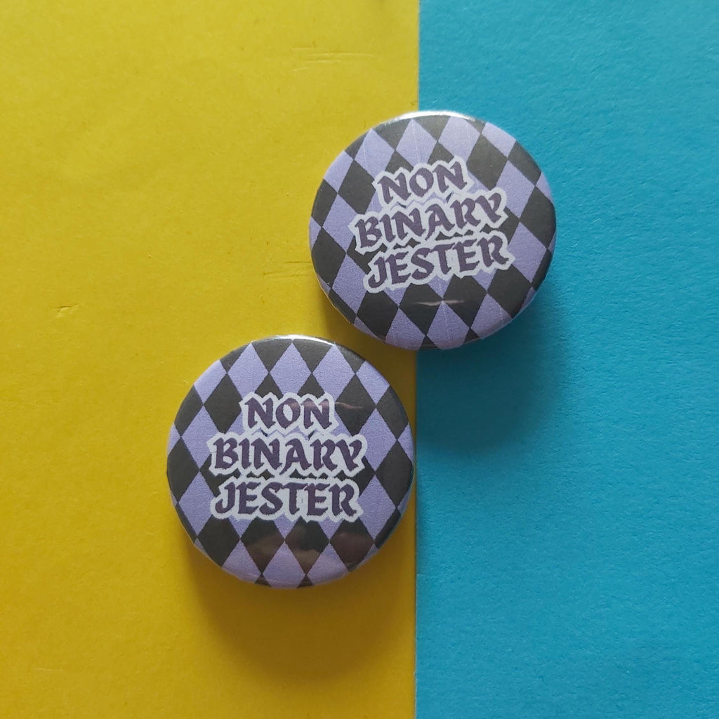 Two round pin-back buttons with a black and lavender harlequin pattern and the text 'NON BINARY JESTER' in white, displayed on a split yellow and blue background.