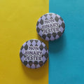 Two round pin-back buttons with a black and lavender harlequin pattern and the text 'NON BINARY JESTER' in white, displayed on a split yellow and blue background.