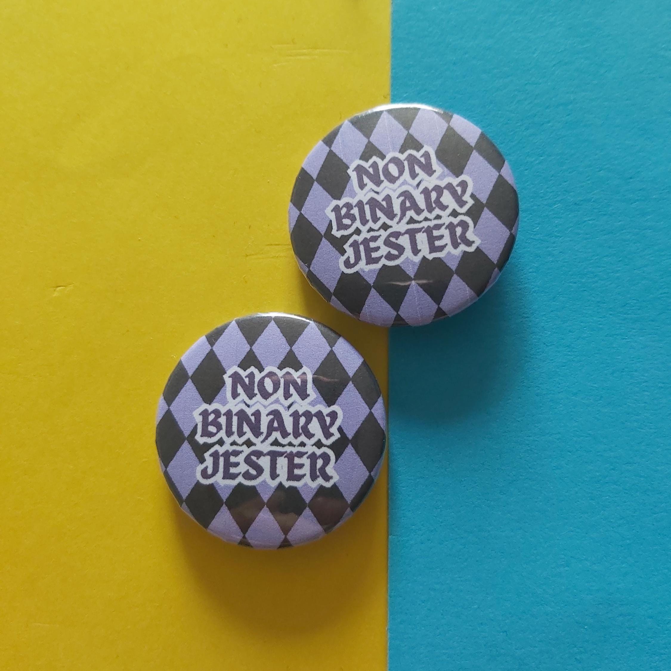 Two round pin-back buttons with a black and lavender harlequin pattern and the text 'NON BINARY JESTER' in white, displayed on a split yellow and blue background.