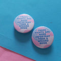 Two round pin-back buttons with the text 'TRANS RIGHTS ARE MORE IMPORTANT THAN A WIZARD BOOK' in blue on a pink and white marbled background, displayed on a blue surface with a pink corner visible.