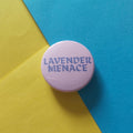 A round pink pinback button badge with the text 'LAVENDER MENACE' in dark purple lettering, set against a diagonally split yellow and blue background.