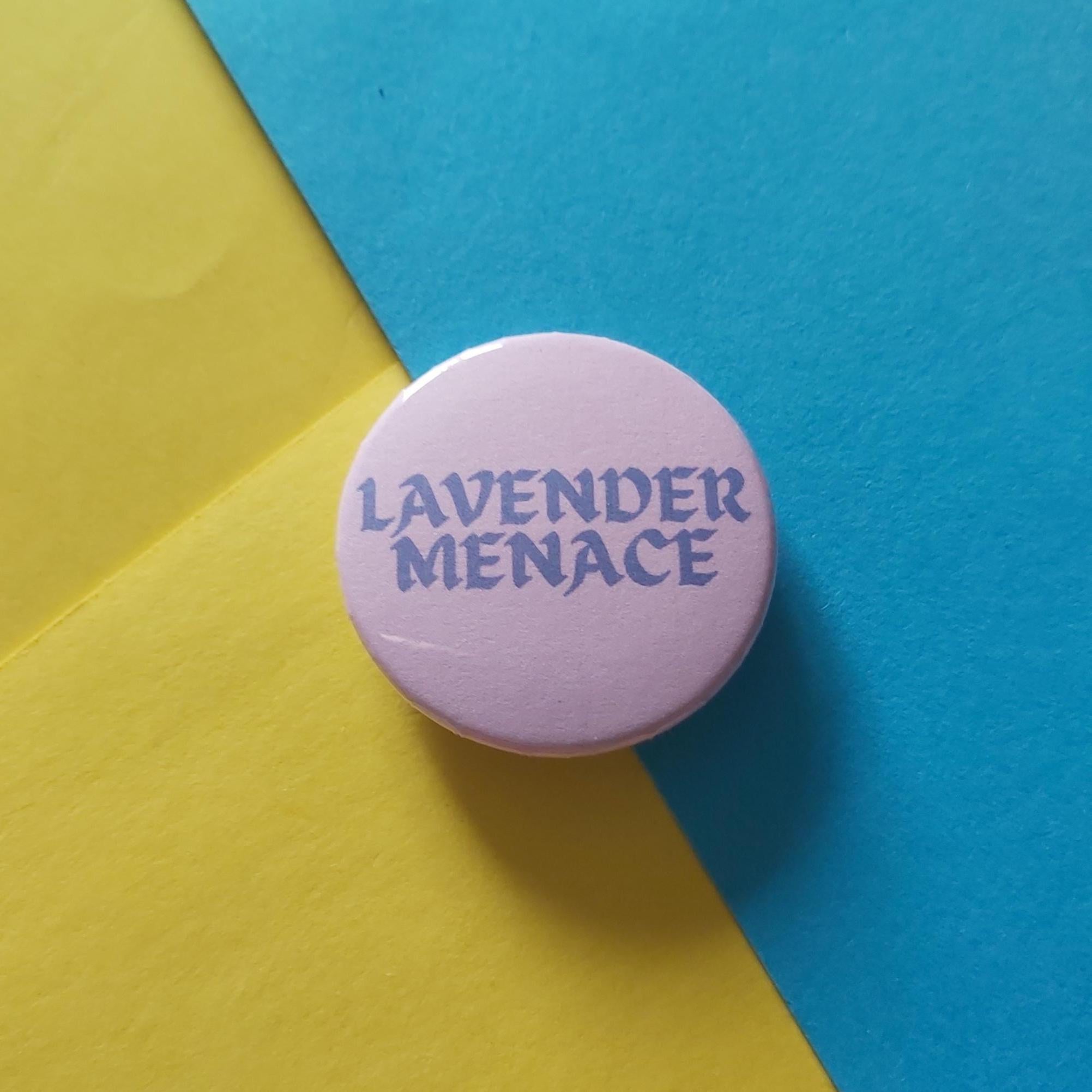 A round pink pinback button badge with the text 'LAVENDER MENACE' in dark purple lettering, set against a diagonally split yellow and blue background.