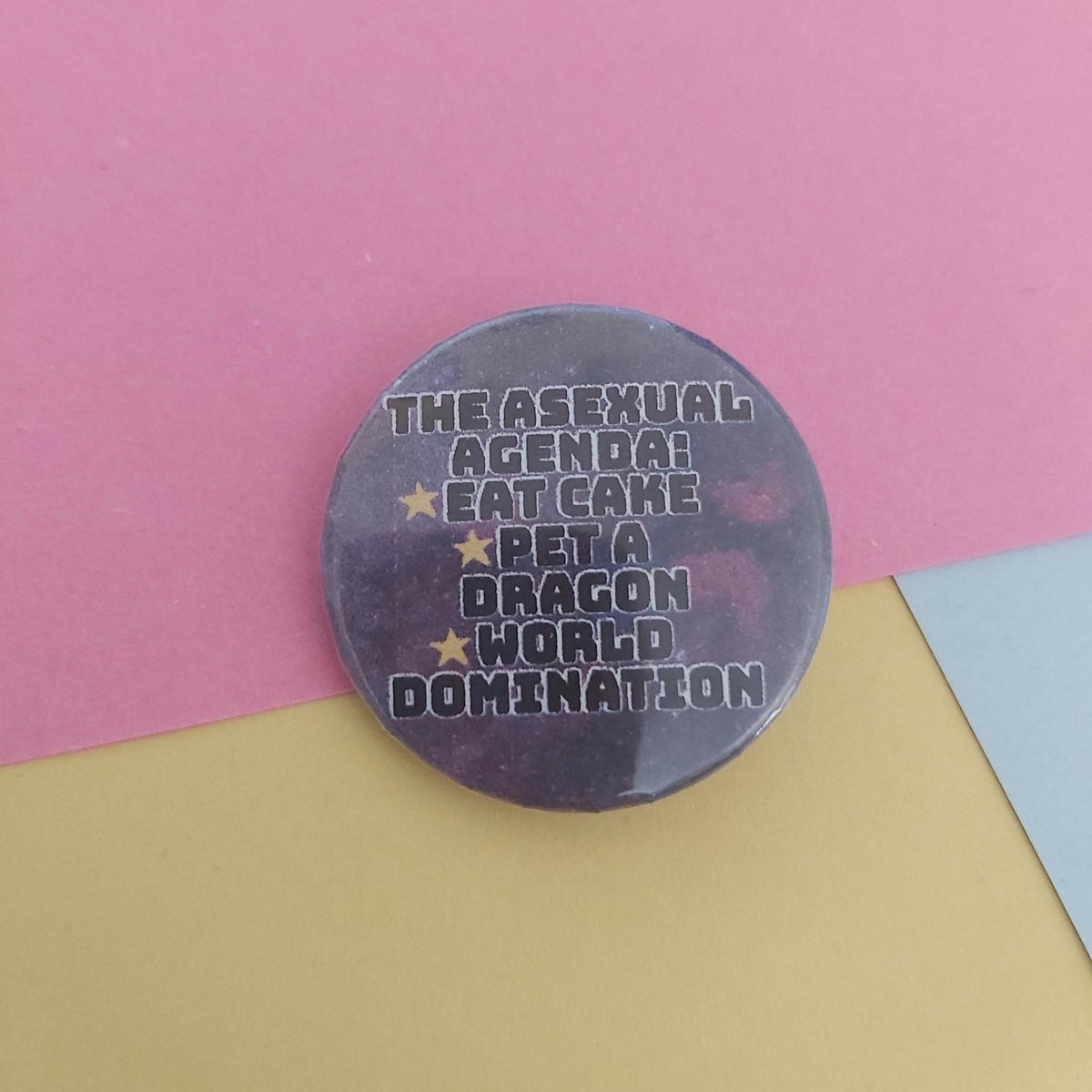 A round pin badge with a dark purple background and white text reading 'THE ASEXUAL AGENDA! EAT CAKE BET A DRAGON WORLD DOMINATION' with small yellow stars, photographed on a pink and yellow paper background.
