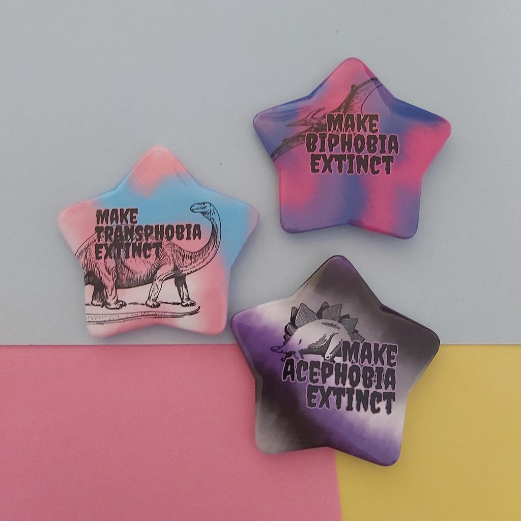 Three star-shaped enamel pins with different pastel designs and text slogans ('MAKE BIPHOBIA EXTINCT', 'MAKE TRANSPHOBIA EXTINCT', 'MAKE ACEPHOBIA EXTINCT')