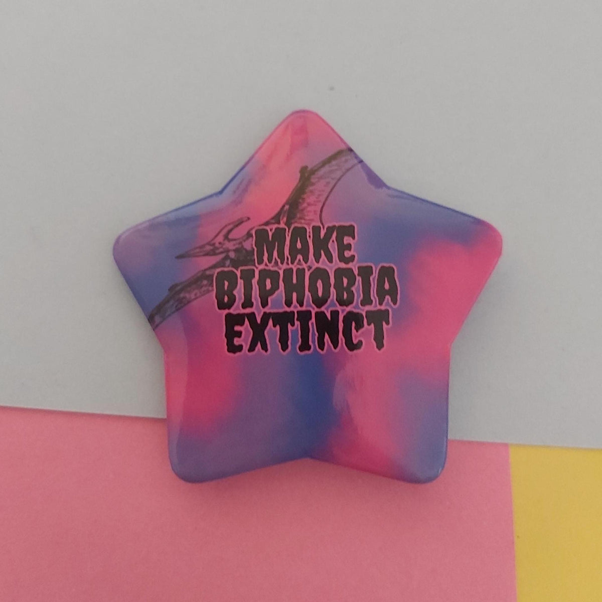 A star-shaped pin with a pink, blue and purple tie-dye pattern, featuring a black pterodactyl graphic and the text 'MAKE BIPHOBIA EXTINCT' in black with a dark pink outline.