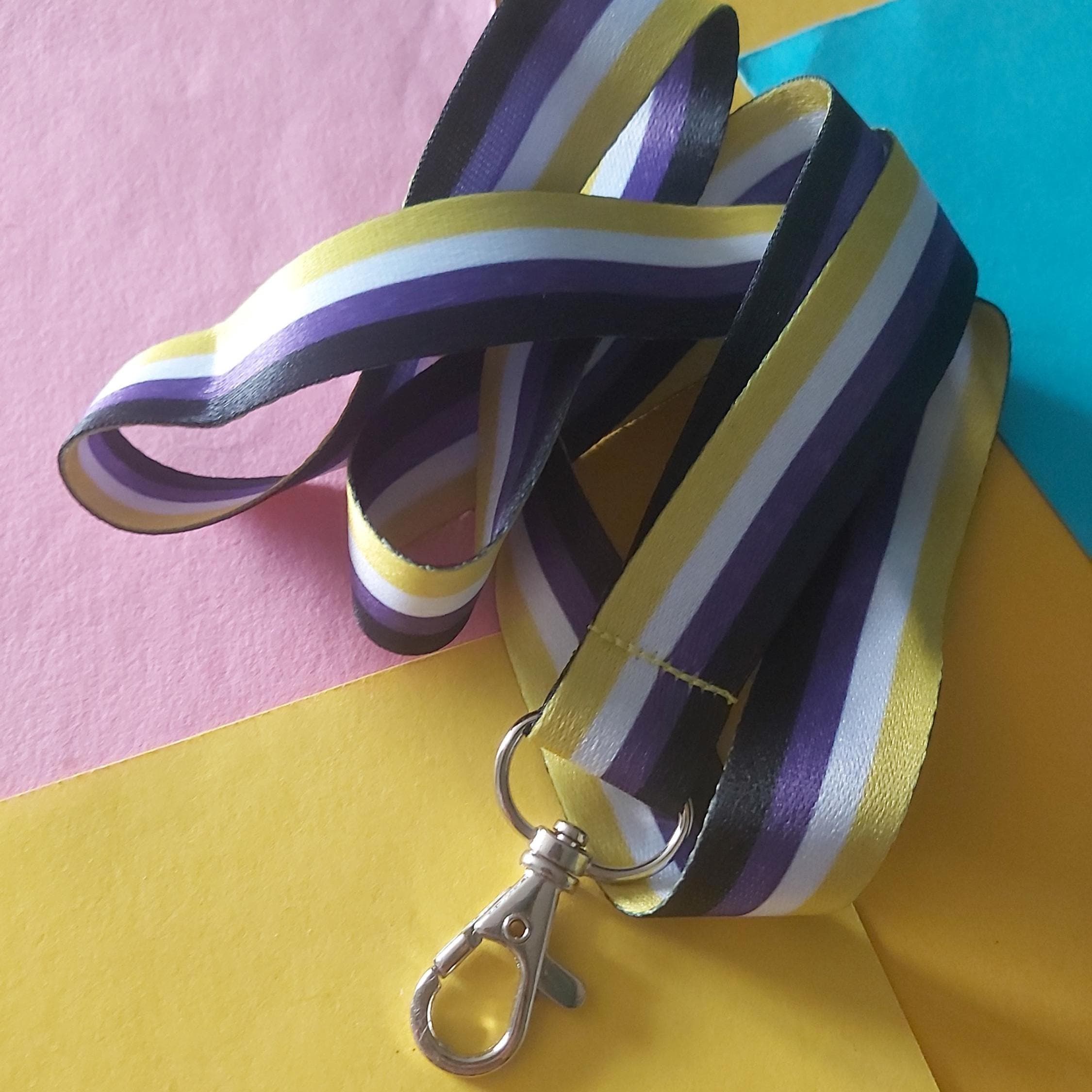 A striped lanyard with a silver lobster clasp, featuring purple, yellow, white, and black stripes of the non-binary flag, laid out on a colourful background of pink, yellow, and teal paper.