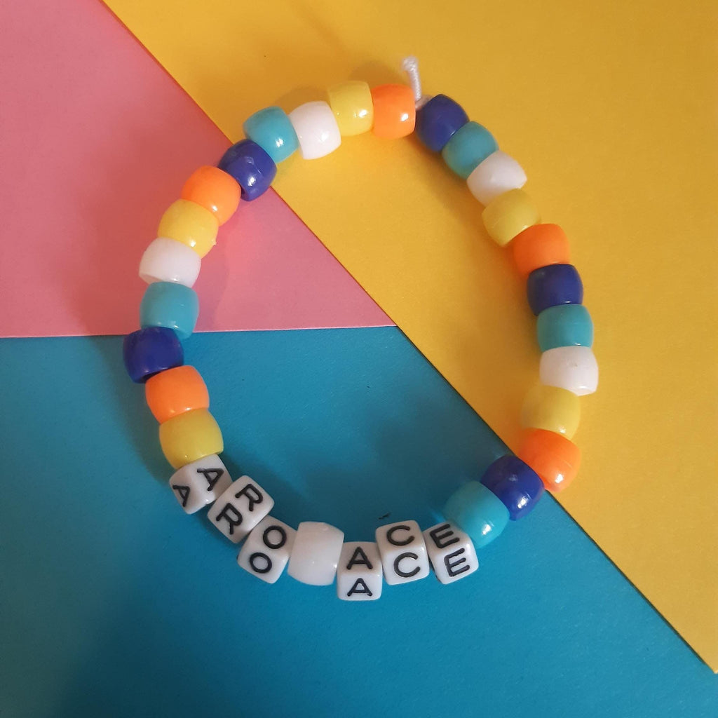 A colourful beaded bracelet with alternating pastel yellow, orange, blue, and white beads, plus white letter beads spelling 'ARO ACE', photographed on a bright multicolour paper background.