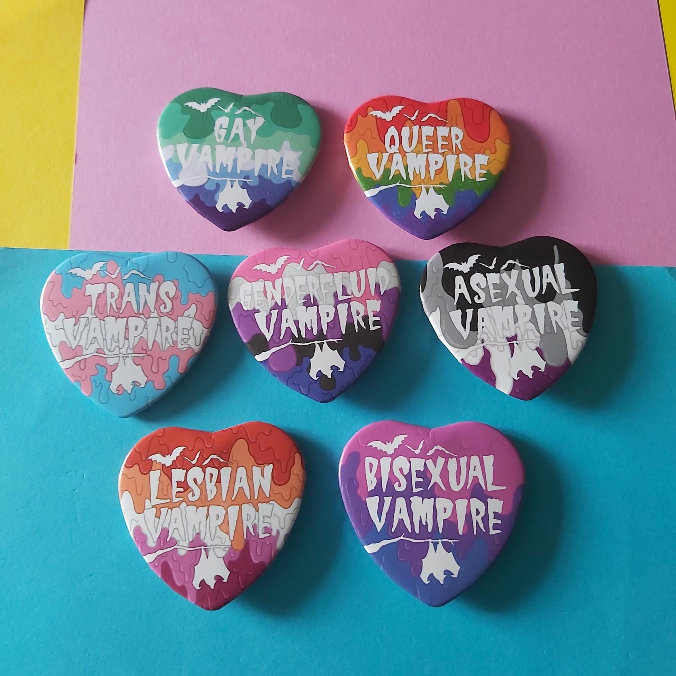 Six heart-shaped pinback buttons featuring LGBTQ+ identity and vampire themes, arranged on a two-tone pink and teal background. The buttons display various pride flags in a paint drip design with white text reading 'GAY VAMPIRE', 'QUEER VAMPIRE', 'TRANS VAMPIRE', 'BISexual VAMPIRE', 'ASexual VAMPIRE', and 'LESBIAN VAMPIRE'.