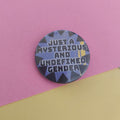 A round pin badge with the text 'JUST A MYSTERIOUS AND UNDEFINED GENDER' in white over a blue, black, and yellow geometric pattern, set against a split pink and yellow background.