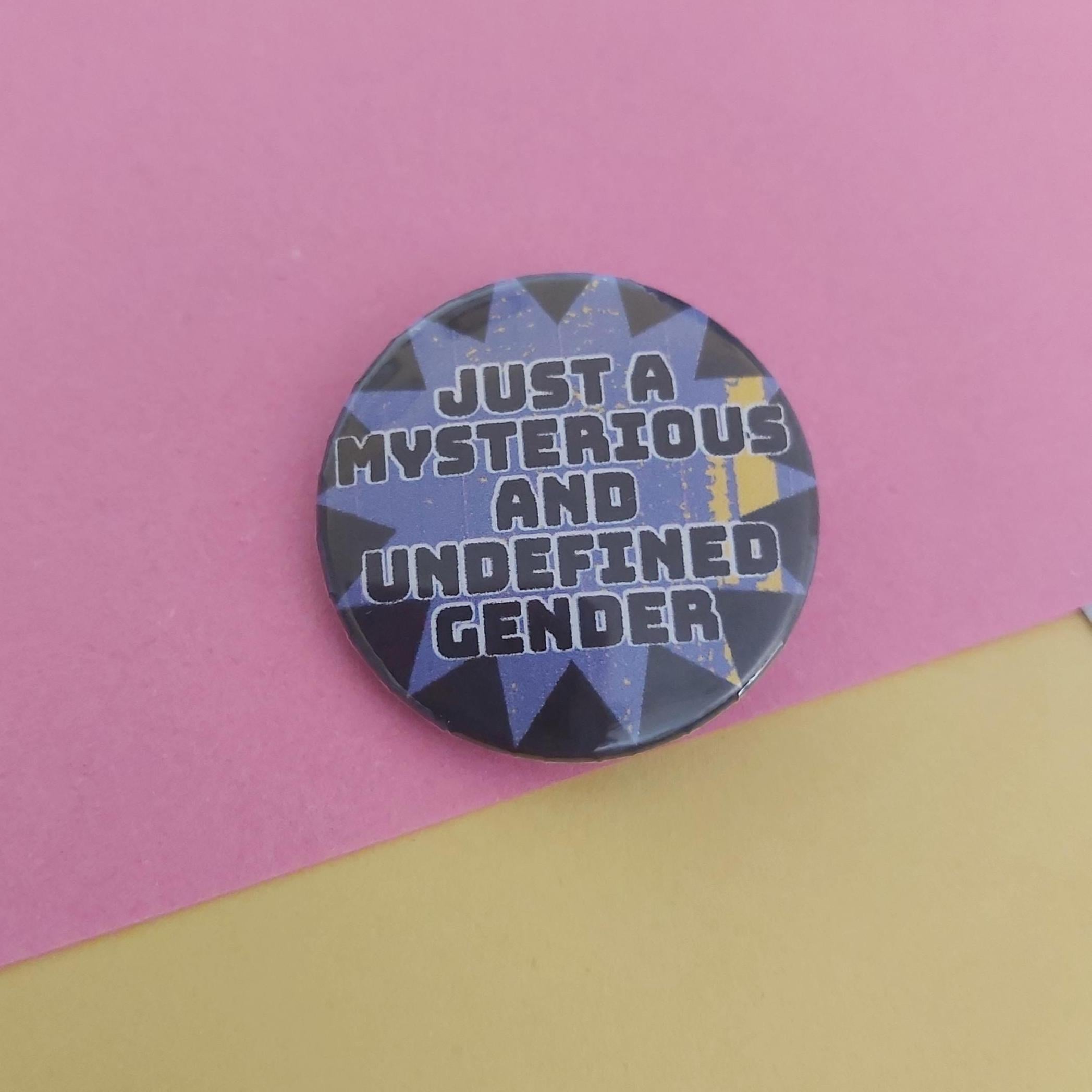 A round pin badge with the text 'JUST A MYSTERIOUS AND UNDEFINED GENDER' in white over a blue, black, and yellow geometric pattern, set against a split pink and yellow background.