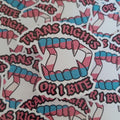 A close-up of multiple die-cut stickers featuring a vampire fang design with pink and blue stripes, the words “TRANS RIGHTS OR I BITE” below, arranged on a white background.