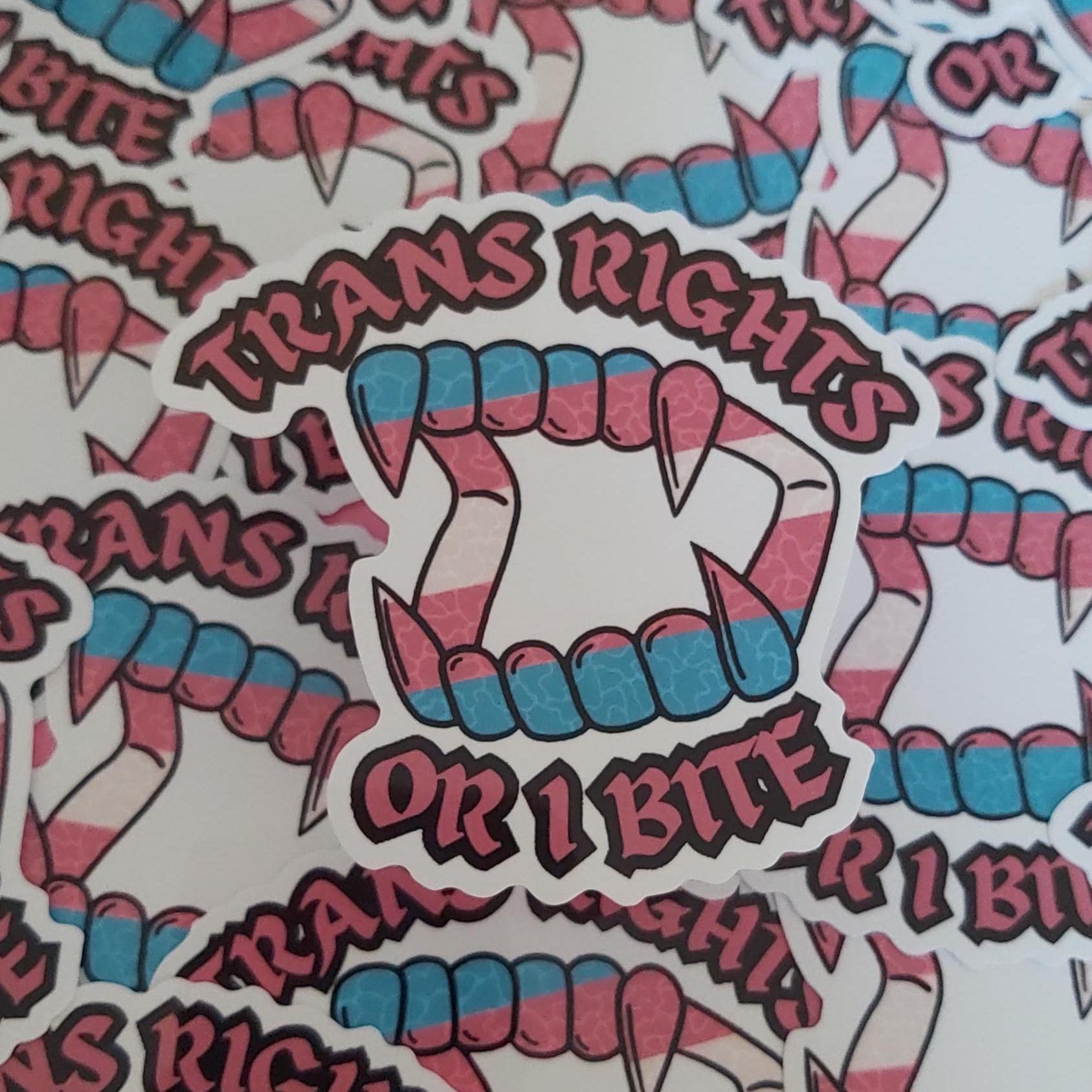 A close-up of multiple die-cut stickers featuring a vampire fang design with pink and blue stripes, the words “TRANS RIGHTS OR I BITE” below, arranged on a white background.