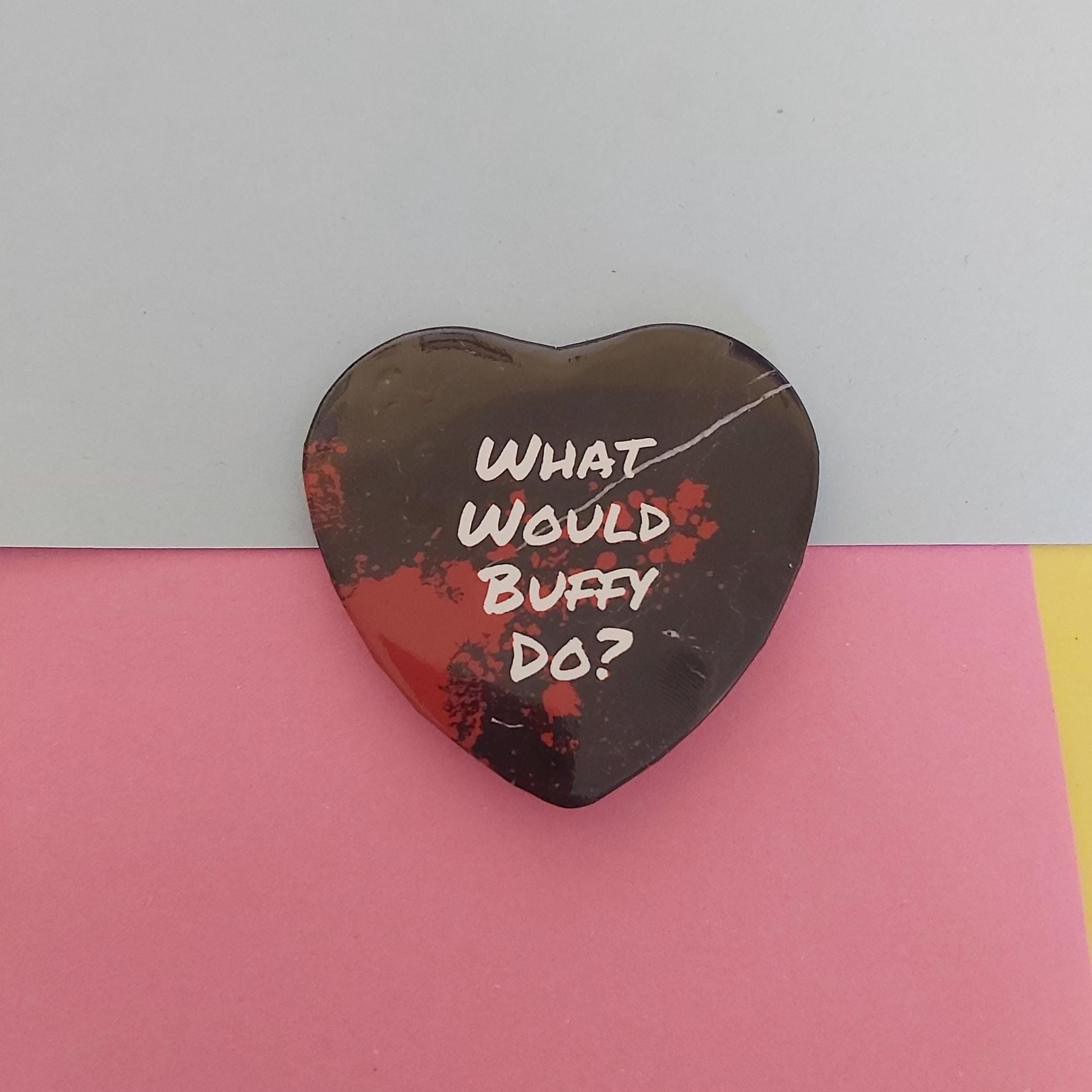 A heart-shaped pin with a black background and red splatter design, featuring the white handwritten text 'WHAT WOULD BUFFY DO?' on a split pink and light blue background.