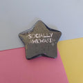 A star-shaped pin with the text 'SOCIALLY AWKWARD' and small sparkle graphics, displayed on a background of pink, yellow, and light blue paper.