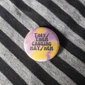 A round pin badge with a purple and yellow tie-dye pattern and the text 'THEY/ THEM CAUSING MAY/HEM' in black, resting on a black and grey striped fabric background.