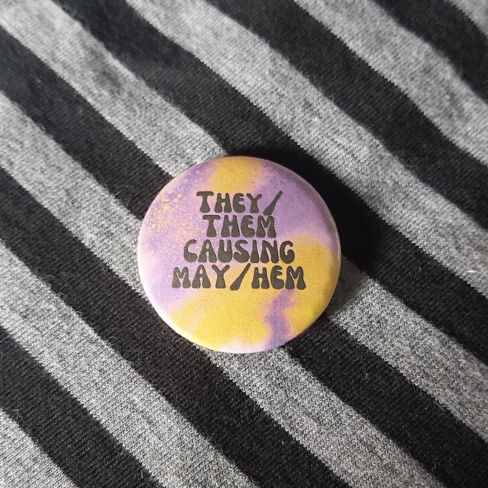 A round pin badge with a purple and yellow tie-dye pattern and the text 'THEY/ THEM CAUSING MAY/HEM' in black, resting on a black and grey striped fabric background.