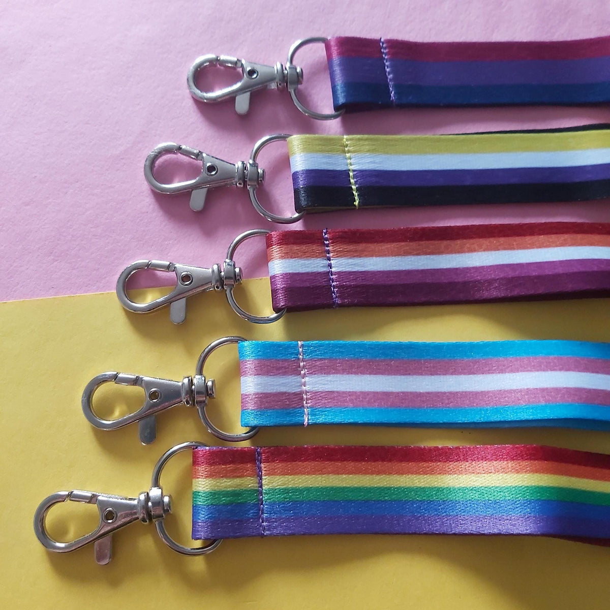 Five cotton Pride flag striped lanyards with silver lobster clasp attachments, arranged diagonally on a split pink and yellow background.