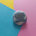 A round pin badge with a colourful marbled pattern in blue, pink, and yellow, featuring the text 'I AM NOT A MISS OR MISTER. I AM A MYSTERY' in dark grey.