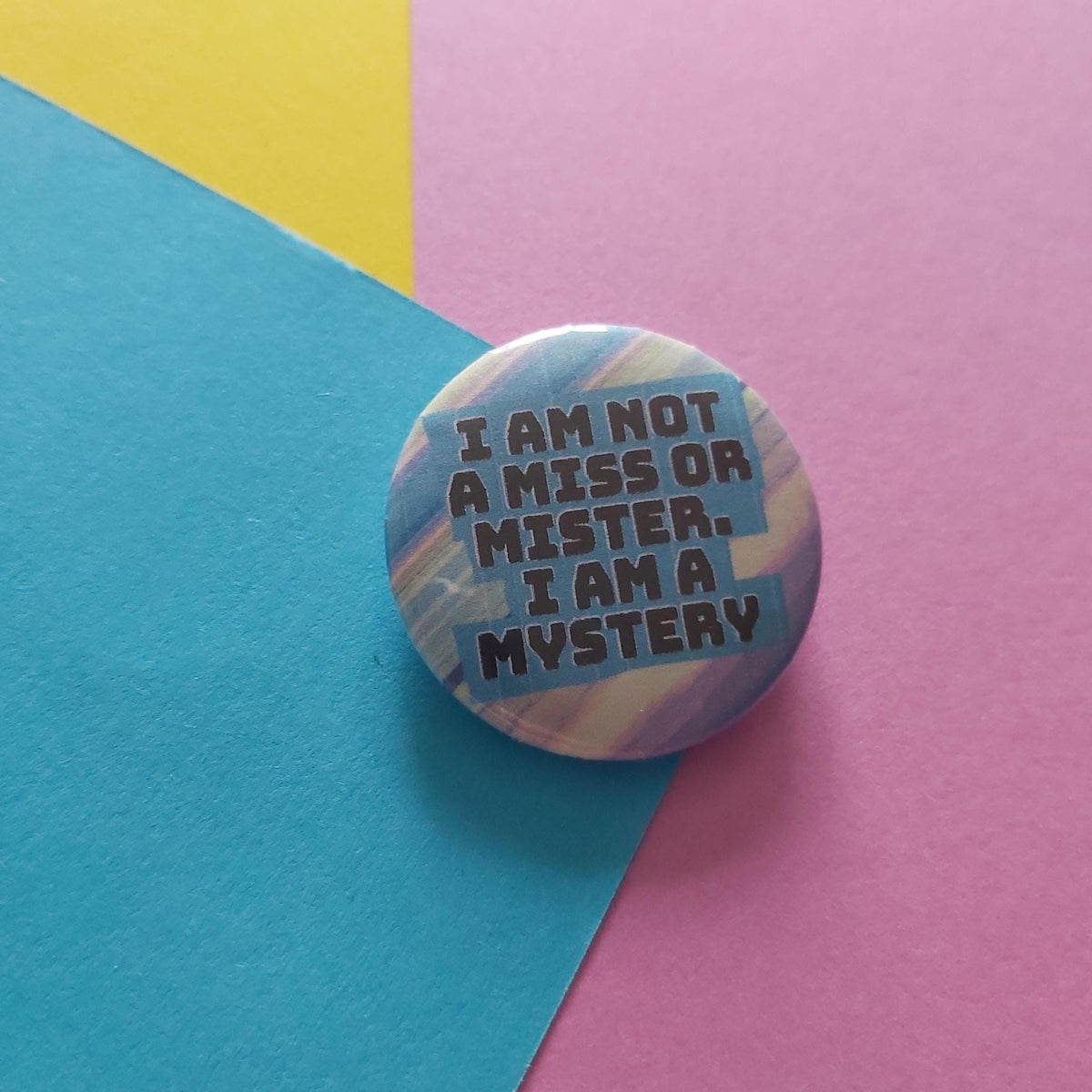 A round pin badge with a colourful marbled pattern in blue, pink, and yellow, featuring the text 'I AM NOT A MISS OR MISTER. I AM A MYSTERY' in dark grey.