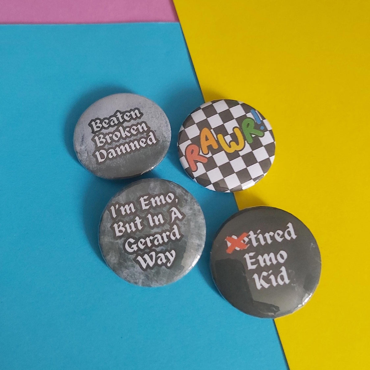 Four round pin-back buttons arranged on a bright blue and yellow background. The buttons have grey, black-and-white checkered, and metallic finishes with text including 'Beaten Broken Damned,' 'I'm Emo But In A Gerard Way,' 'RAWR,' and 'Retired Emo Kid.