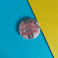 A round pin-back button badge featuring the words 'Sword Wielding Sapphic' in pink gothic lettering over a dark sword photograph, placed on a diagonally split blue and yellow background.