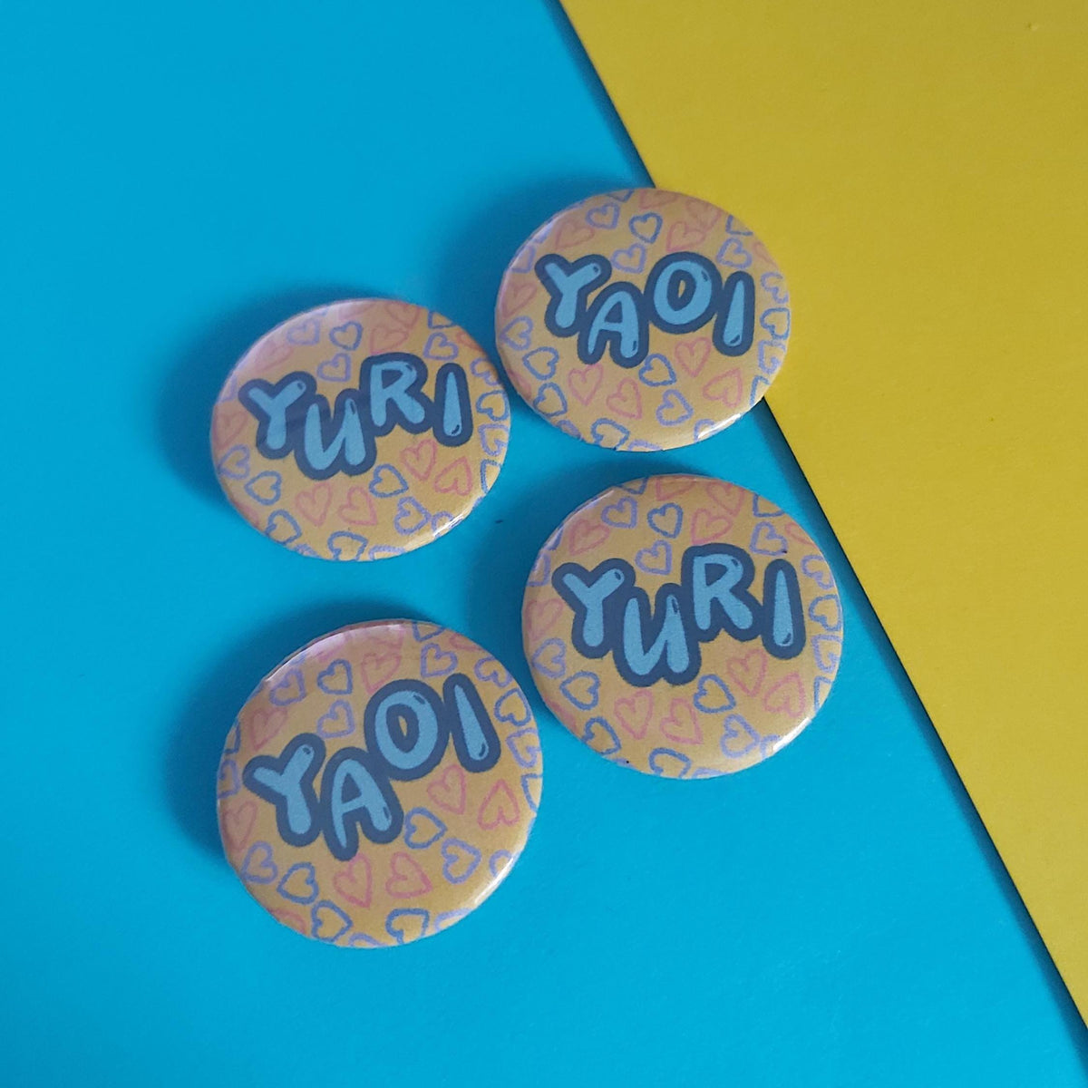Four round pinback buttons featuring the words 'YURI' and 'YAOI' in blue bubble letters over a peach and pink patterned background, arranged on a diagonally split blue and yellow surface.