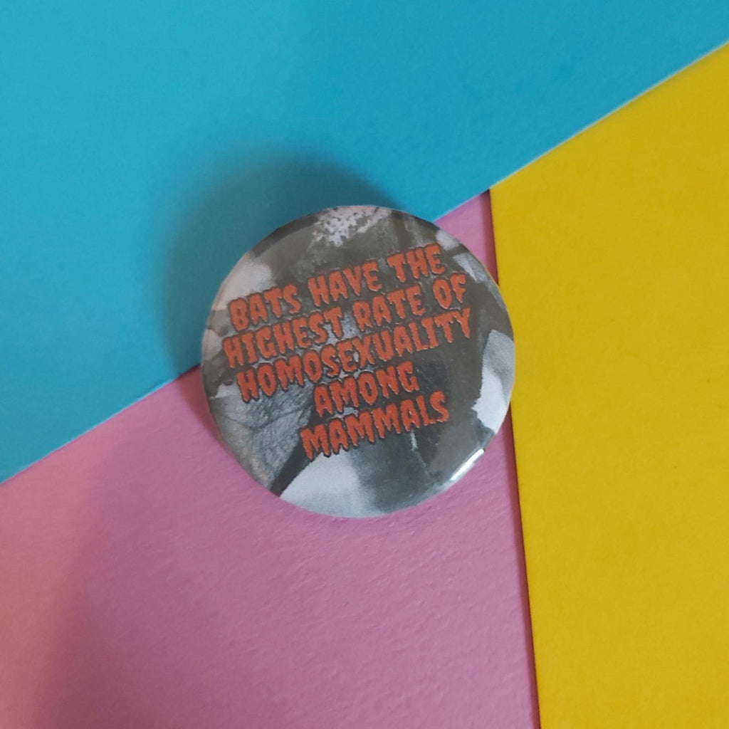 A round pinback button badge featuring a grayscale bat photo with red text that reads, 'BATS HAVE THE HIGHEST RATE OF HOMOSEXUALITY AMONG MAMMALS,' placed on a colourful background of cyan, pink, and yellow paper.