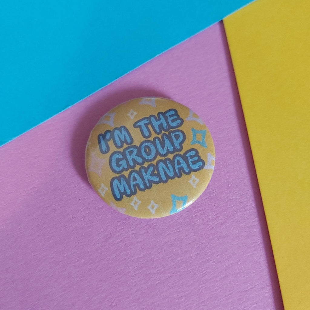 A round pinback button badge with the text 'I'M THE GROUP MAKNAE' in blue letters on a yellow background with small white and blue star patterns, resting on a pink surface with diagonal blue and yellow paper borders.