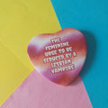 A heart-shaped badge with a pink and orange tie-dye pattern and white text reading 'THE FEMININE URGE TO BE SEDUCED BY A LESBIAN VAMPIRE', placed on a diagonally split yellow, blue, and pink background.