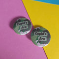 Two round pinback buttons with a green camouflage pattern and the text 'Cupid's Aro' in white, displayed on a diagonally split pink and yellow paper background.