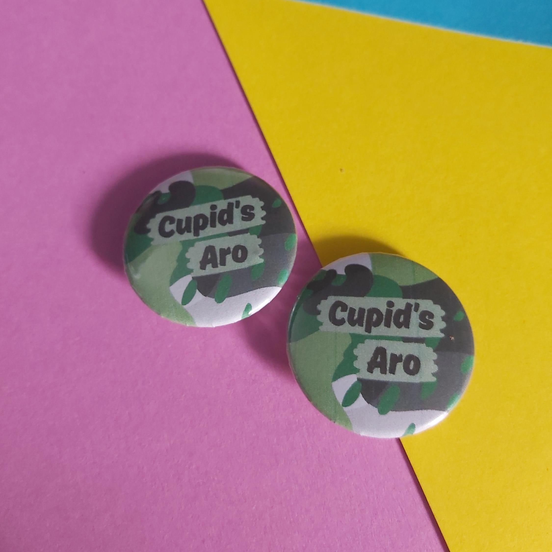 Two round pinback buttons with a green camouflage pattern and the text 'Cupid's Aro' in white, displayed on a diagonally split pink and yellow paper background.