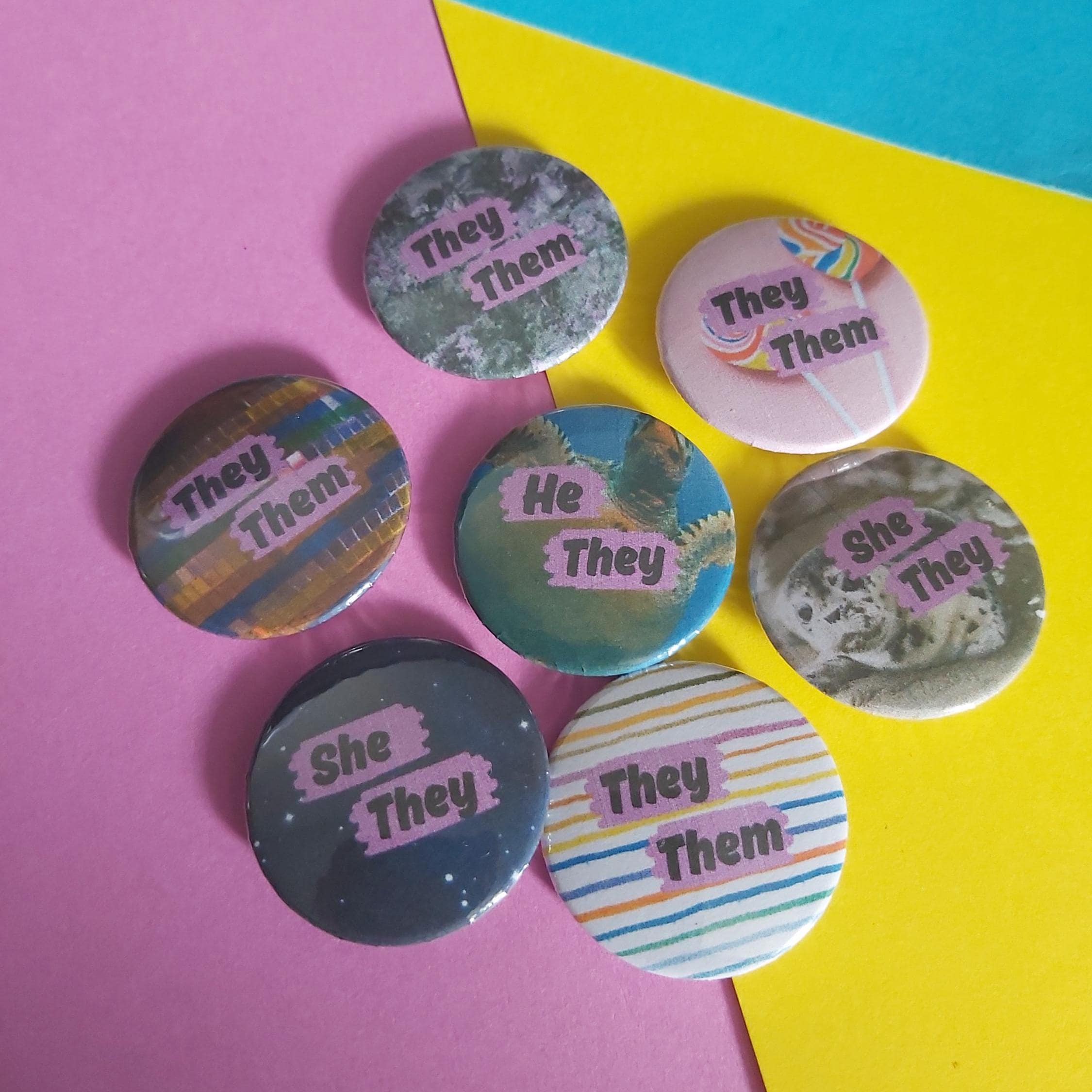 Seven round pinback buttons with various printed designs and the text 'They She They' or 'She They' in pink, arranged on a diagonally split pink and yellow background.