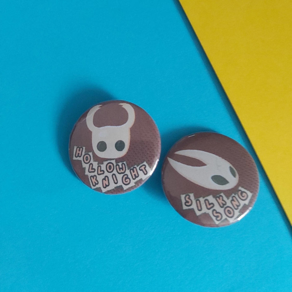 Two round pin-back buttons featuring Hollow Knight silhouettes on a textured brown background, placed on a bright blue surface with a yellow corner accent.