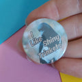 A close-up of a hand holding a round pinback button badge with the text 'I Like Shiny Trinkets' over a dark silhouette on a light background, set against colorful pink, blue, and yellow paper.