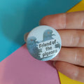 A close-up of a hand holding a round pinback button badge with a light blue background, featuring two dark blue-grey pigeons and the text 'Friend of the pigeons' in black. The background consists of colorful paper sheets in teal, pink, and yellow.