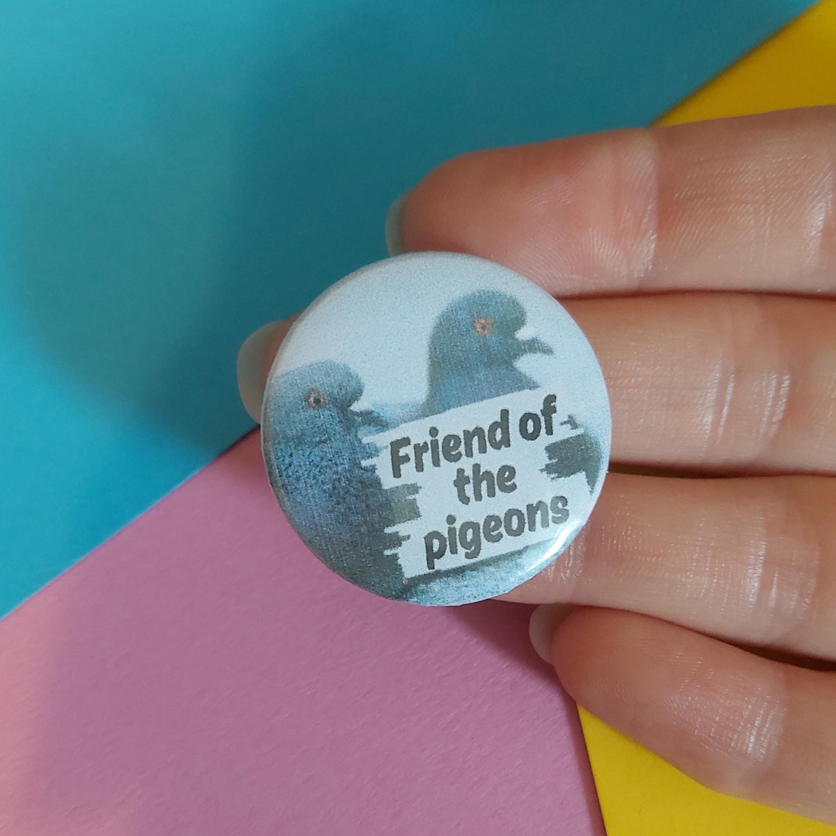 A close-up of a hand holding a round pinback button badge with a light blue background, featuring two dark blue-grey pigeons and the text 'Friend of the pigeons' in black. The background consists of colorful paper sheets in teal, pink, and yellow.