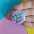 A close-up of a hand holding a round pinback button badge with the text 'Putting the Ace in Menace' in black lettering on a light purple background with small white star details.