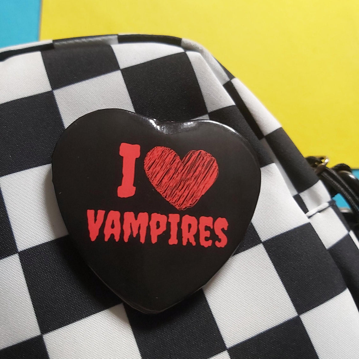 A black heart-shaped pin with red text reading 'I ♥ VAMPIRES' is attached to a black-and-white checkered bag, set against a bright yellow background.