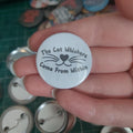 A hand holding a round pin-back button badge with the text 'The Cat Whiskers Come From Within' and a simple whisker-and-heart nose graphic.