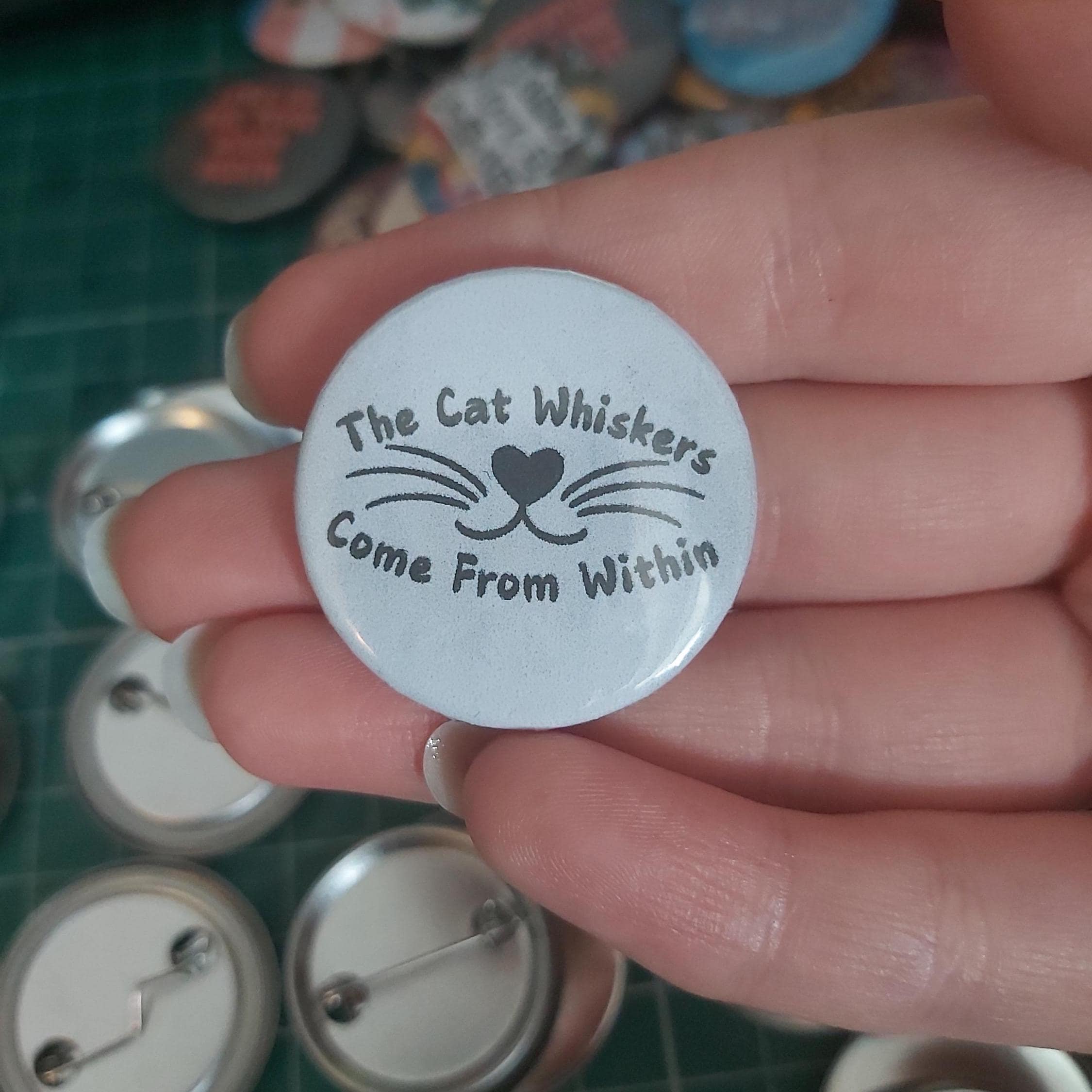 A hand holding a round pin-back button badge with the text 'The Cat Whiskers Come From Within' and a simple whisker-and-heart nose graphic.