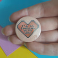 A hand holding a round pinback button badge with a pixelated heart design and the text 'I'm in lesbian with you' in dark lettering on a peach and pink background.