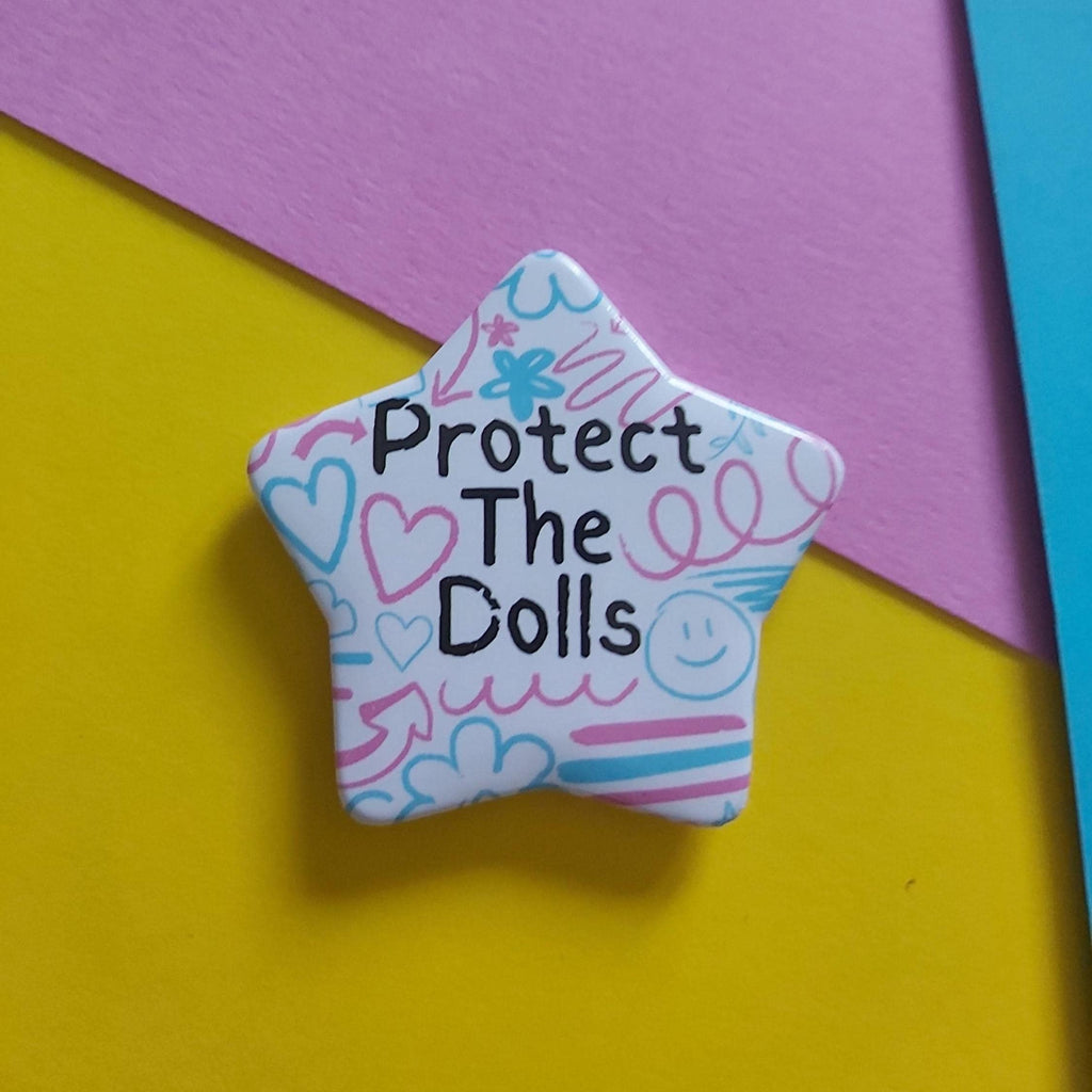 A star-shaped enamel pin with the text 'Protect The Dolls' in black, decorated with pink, blue, and purple doodles on a white background, set against a colorful yellow, pink, and blue paper backdrop.