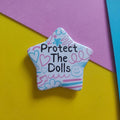 A star-shaped enamel pin with the text 'Protect The Dolls' in black, decorated with pink, blue, and purple doodles on a white background, set against a colorful yellow, pink, and blue paper backdrop.