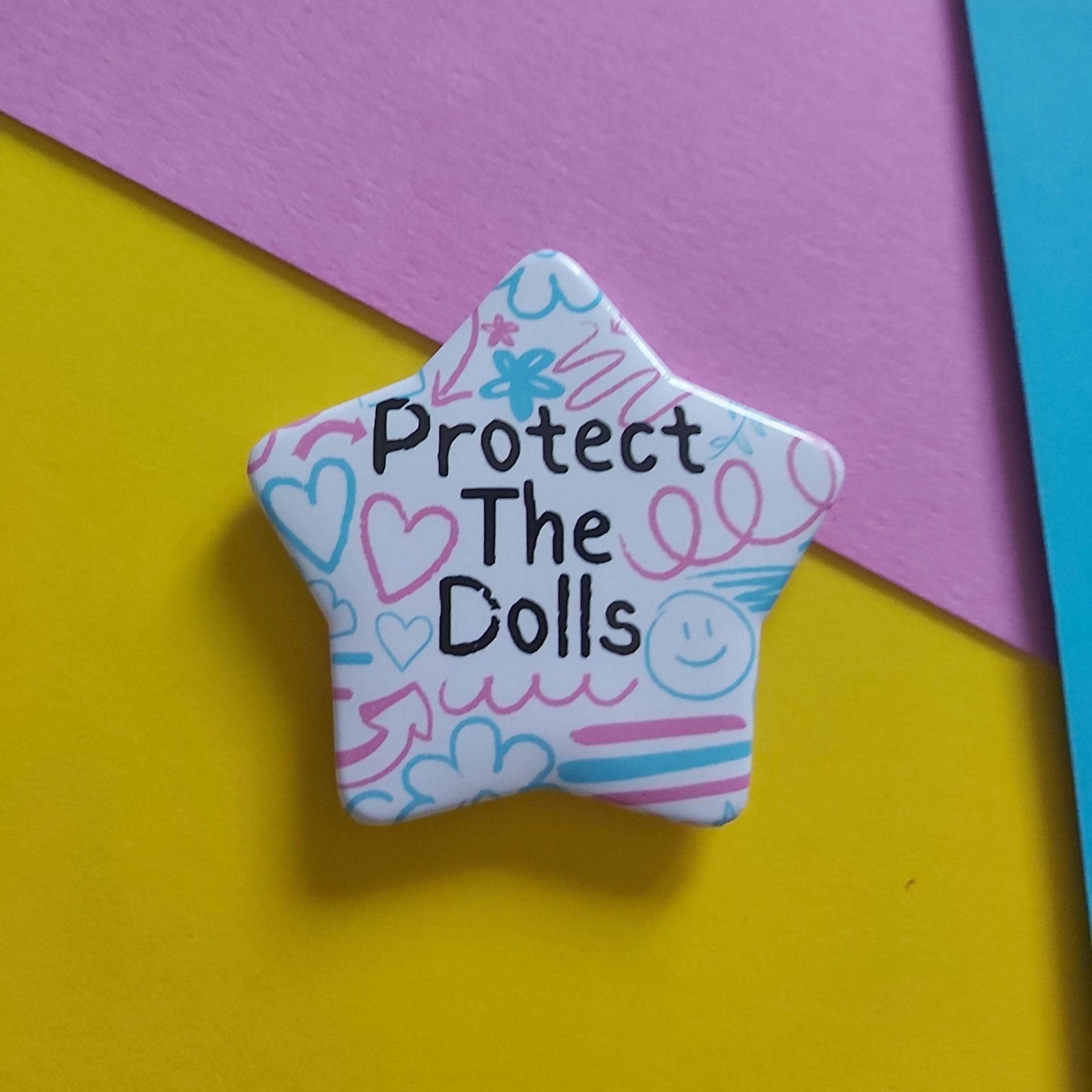 A star-shaped enamel pin with the text 'Protect The Dolls' in black, decorated with pink, blue, and purple doodles on a white background, set against a colorful yellow, pink, and blue paper backdrop.