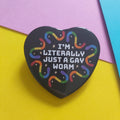 A heart-shaped black pin with the text 'I'M LITERALLY JUST A GAY WORM' in white, surrounded by a rainbow-colored gummy worms and small stars, displayed on a colourful background of yellow, pink, and blue paper.