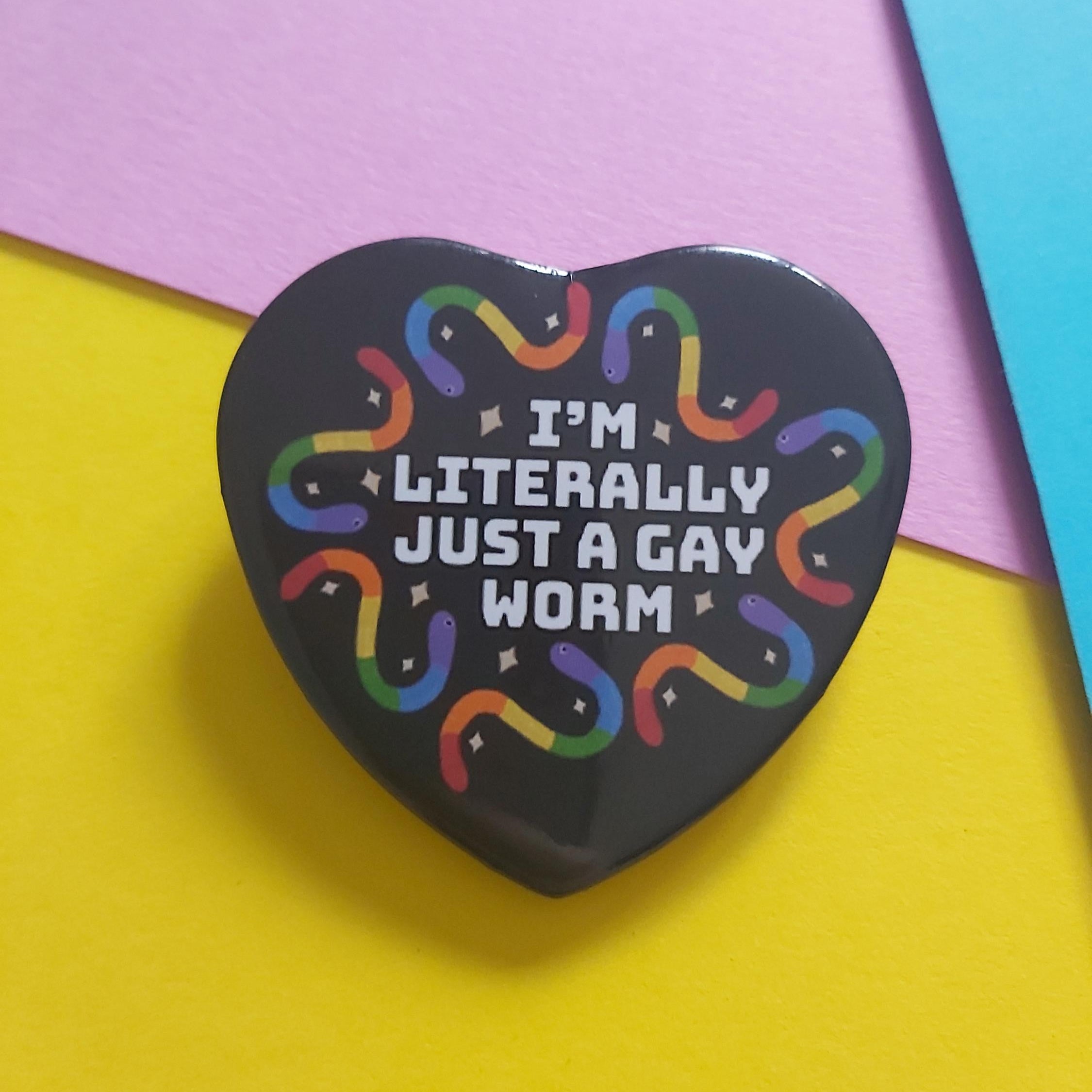 A heart-shaped black pin with the text 'I'M LITERALLY JUST A GAY WORM' in white, surrounded by a rainbow-colored gummy worms and small stars, displayed on a colourful background of yellow, pink, and blue paper.
