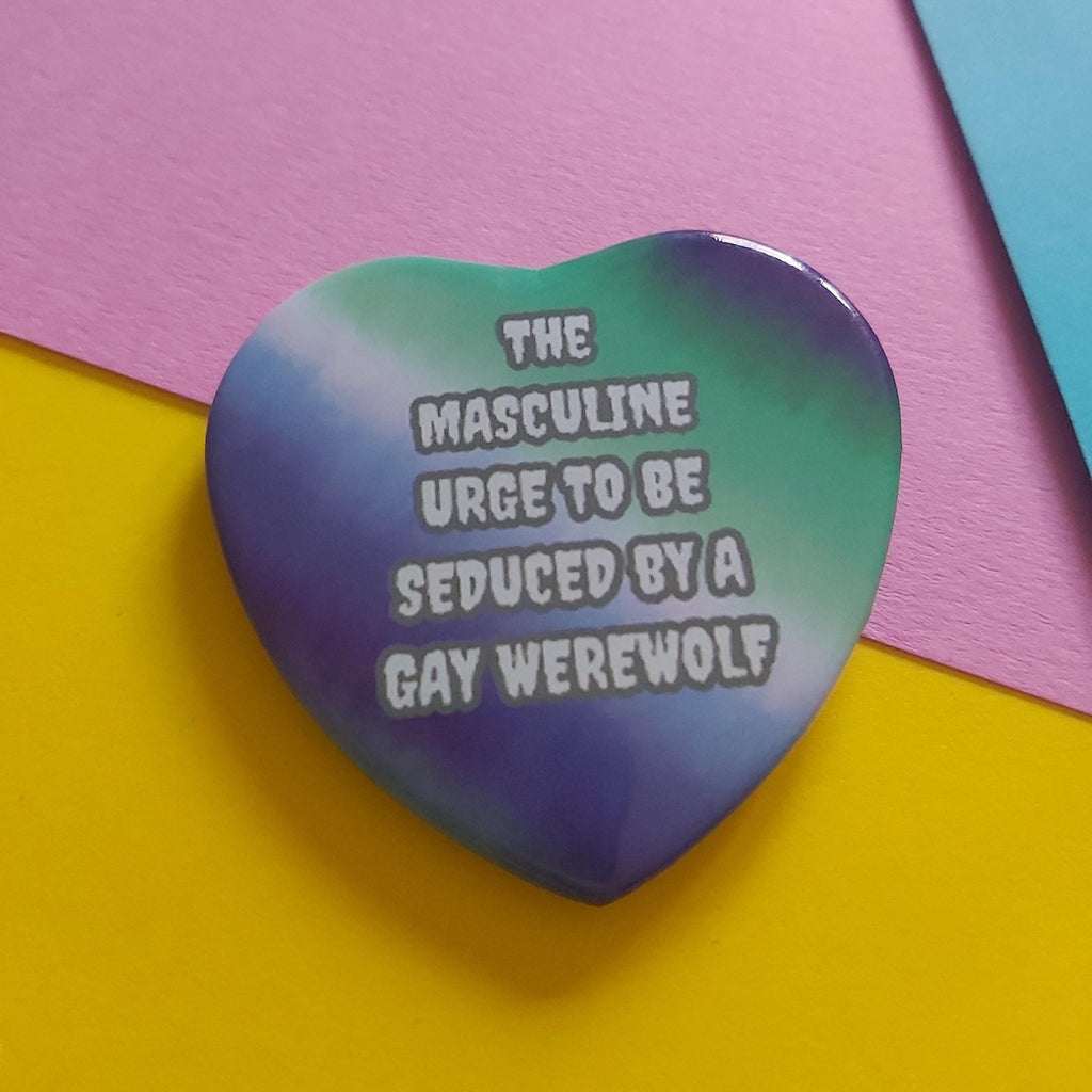 A heart-shaped pinback button badge featuring a green and teal gradient background with the text 'THE MASCULINE URGE TO BE SEDUCED BY A GAY WEREWOLF' in white with a black outline. The badge is displayed on a colourful background of pink, yellow, and blue paper.