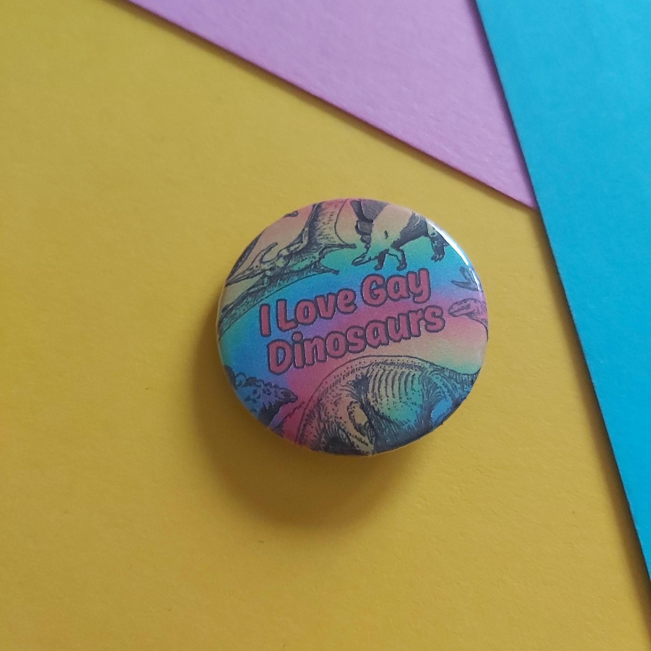 A round pinback button badge featuring a colourful dinosaur illustration and the text 'I Love Gay Dinosaurs' in red, placed on a yellow background with purple and blue paper accents.