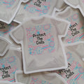A die-cut sticker shaped like a short-sleeve T-shirt, featuring the text 'Protect The Dolls' in black alongside pink and blue retro doodles, displayed on a pile of similar stickers.